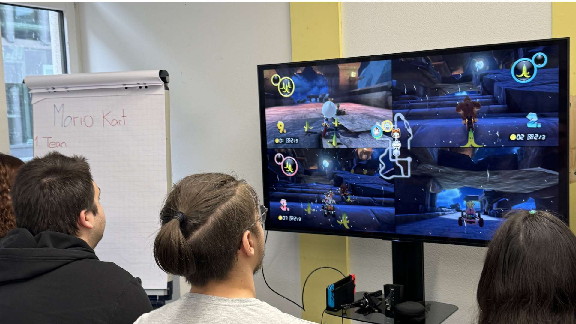 Vier Klar.Doch. Mitarbeitenden spielen Mario Kart auf einem grossen Fernseher. Ein Flipchart zeigt die Spielstände nebenan. Es herrscht eine lockere und freundschaftliche Atmosphäre.